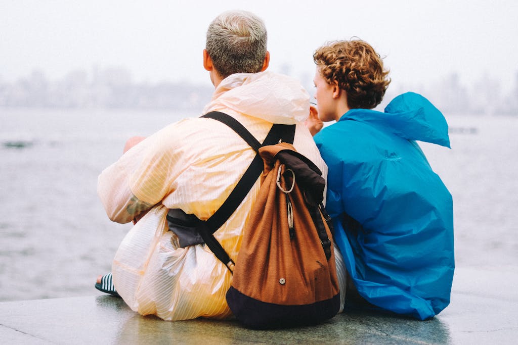 Man and Teenage Boy in Raincoats Sitting by a Sea
